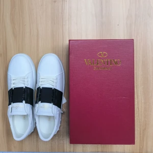 Valentino Garavani Open sneakers vit/svart - Snygga Valentino Garavani Open sneakers i vitt skinn med bred svart rem över vristen. Klassisk rund tå, platt sula med nitar baktill och diskret logga på plösen. Perfekta för dig som gillar stilrena och lyxiga detaljer.