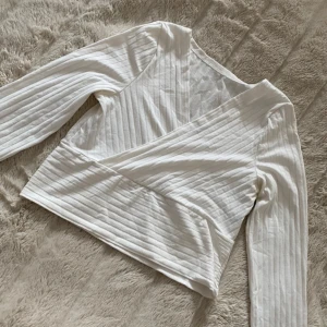 Vit omlottopp med ribbade detaljer - Snygg vit omlottopp med långa ärmar och ribbad struktur. Toppen har en croppad passform och omlottdesign som ger en modern och fräsch look. Perfekt att matcha med höga jeans eller kjol för en trendig outfit.