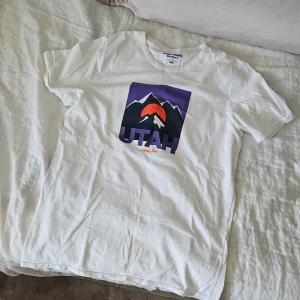 Vit t-shirt med Utah-tryck Jack & Jones - Vit t-shirt från Jack & Jones med coolt grafiskt tryck av berg, solnedgång och texten 'UTAH' i lila och orange på bröstet. Klassisk passform och rund halsringning, perfekt till jeans eller shorts. Skön bomullskvalitet.