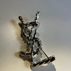 Saint Laurent silver brosch - Säljer en unik brosch från Saint Laurent i blankt silverfärgat metall. Broschen har YSL-loggan i en vriden, rep-liknande design som ger en cool och edgy vibe. Perfekt accessoar för att lyfta vilken outfit som helst.
