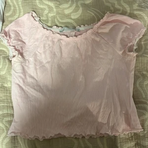 Ljusrosa blus med volangkant - Supersöt ljusrosa blus med korta ärmar och volangkant vid halsringning och ärmslut. Tunt, lätt material med lite skrynklig struktur som ger en chill och romantisk vibe. Perfekt att styla med jeans eller kjol för en söt look.