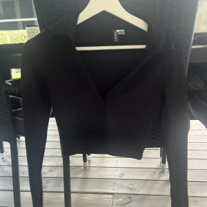 Svart ribbad croppad topp H&M Divided - Svart croppad topp från H&M Divided med lång ärm och djup v-ringning. Toppen är ribbad och har en klassisk krage som ger en snygg och trendig look. Perfekt att styla med höga jeans eller kjol.