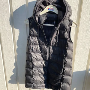 Svart dunväst med huva från Zara - Snygg svart dunväst från Zara med quiltad design och dragkedja framtill. Västen har en stor huva med dragsko och är perfekt för lager-på-lager-stil. Passar dig som gillar en clean och sportig look.