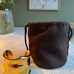 ANI bredt guldarmband i vågig form - Säljer mitt armband från ANI i blankt guldigt metallmaterial. Armbandet har en bred, vågig och organisk form som ger en cool och modern look. Kommer med original brun sammetsförvaringspåse. Aldrig använt 