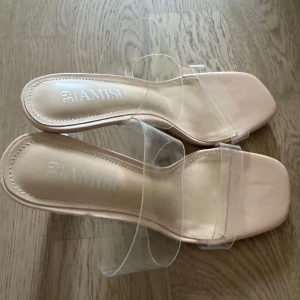 Beiga klackar med transparent rem från Amisu - Snygga beiga klackar från Amisu med transparenta remmar och fyrkantig tå. Skorna har en hög, genomskinlig klack och platt sula framtill. Perfekta för dig som gillar trendiga och stilrena detaljer.