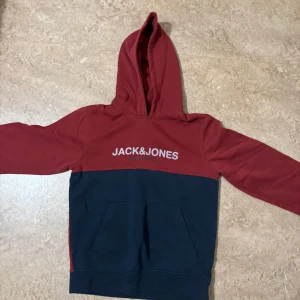 Röd och marinblå hoodie Jack & Jones - Snygg hoodie från Jack & Jones i rött och marinblått med stor logga framtill. Tröjan har huva, känguruficka och långa ärmar. Perfekt för chill dagar eller när du vill ha en avslappnad stil. Mjuk insida i bomullsmix för extra komfort.