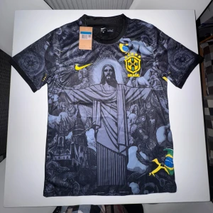 Brasilien x Nike fotbollströja  - Snygg Brasilien fotbollströja från Nike med unikt svart och grått mönster, gult Nike-märke och Brasilien-emblem på bröstet. Tryck av Kristusstatyn och brasilianska flaggan på framsidan. Tillverkad i Dri-FIT-material som andas.
