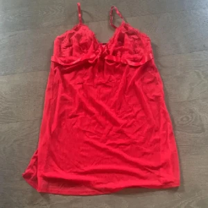 Röd kort nattlinne med rosett - Snygg röd kortklänning i transparent material med spets upptill och smala axelband. Klänningen har en söt rosett framtill och satindetaljer längs bysten. Perfekt för dig som vill sticka ut med en feminin och flirtig look. Tryck ej köp nu