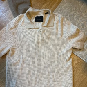 Beige pikétröja från Jack & Jones - Säljer en beige pikétröja från Jack & Jones Premium i storlek M. Tröjan har klassisk krage, korta ärmar och är tillverkad i ett strukturerat, stickat material som ger en snygg och modern look. Perfekt för dig som gillar stilrena och enkla plagg.