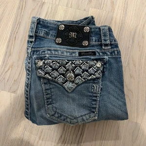 Miss Me bootcut jeans med paljettdetaljer - Snygga blå jeans från Miss Me med bootcut passform. Bakfickorna har grymma paljettdetaljer och silvriga knappar som verkligen sticker ut. Klassisk ljusblå tvätt och coola kontrastsömmar. Perfekta för dig som vill ha jeans med extra attityd. Längd: 103cm Midja 36-38cm (stretch) Benöppning 21cm inseam 81cm