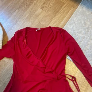 Röd omlottklänning från Gina Tricot - Snygg röd omlottklänning från Gina Tricot med långa ärmar och volangdetalj längs v-ringningen. Klänningen har knyt i midjan och är perfekt för dig som vill sticka ut. Mjuk och följsam känsla, passar dig som gillar trendiga och feminina plagg.