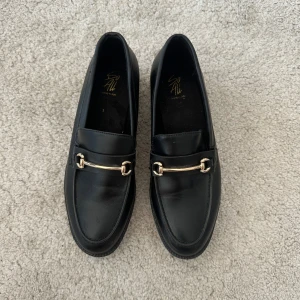 Svarta loafers med gulddetalj - Stilrena svarta loafers med snygg guldfärgad metallspänne över foten. Klassisk design med rund tå och platt sula.