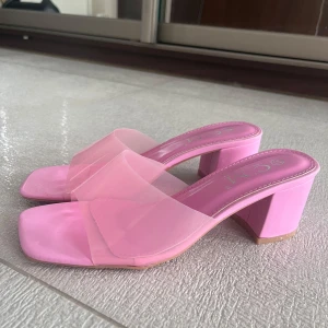 Rosa klackade sandaler med transparent rem - Snygga rosa sandaler med bred transparent rem och fyrkantig tå. Skorna har en stabil blockklack i matchande rosa och en platt sula. De är aldrig använda så nyskick! Nypris: 299kr