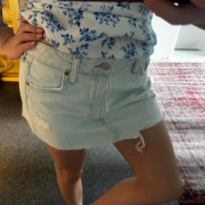 Ljusblå jeansskjol - Säljer en ljusblå jeansskjol med låg midja och rå, fransig kant. Shortsen har klassisk femficksdesign, slitna detaljer och dragkedja med knapp. Perfekta för varma sommardagar och ger en avslappnad vibe.