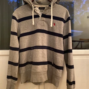 Randig hoodie från Polo Ralph Lauren - Grå hoodie med marinblå ränder från Polo Ralph Lauren. Tröjan har huva med snörning, känguruficka och det klassiska röda Polo-logot broderat på bröstet. Skön bomullskvalitet och långärmad modell, perfekt för chill dagar.