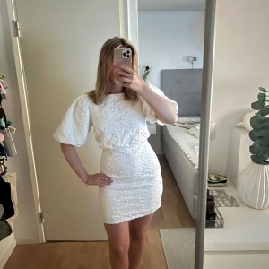 Vit kort klänning med puffärm och rosett - Superfin vit kort klänning med struktur, puffärmar och öppen rygg med stor rosett. Tight passform i midjan och lätt volym upptill. Perfekt för dig som vill sticka ut med en söt och trendig look.