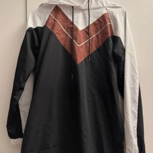 NA-KD klänning med Luva - En typ klänning med material som är likt regnjacke/täckjackematerial. Har ett band som går att knyta både på framsidan och baksidan i midjan.   Är stl 40 (M) men skulle nog även kunna passa stl S.