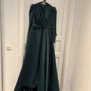 Mörkgrön långklänning med paljetter - Säljer en elegant mörkgrön långklänning med glittriga paljett-detaljer och bälte i midjan. Klänningen har lång ärm, hög hals och ett vackert fall. Perfekt för dig som vill sticka ut med en lyxig och sofistikerad look.
