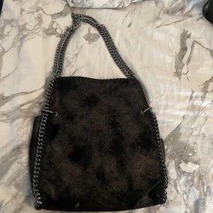 Svart axelväska med kedjedetaljer - Cool svart axelväska med pälsliknande yta och grova silverfärgade kedjor som axelrem. Väskan har en fyrkantig form och insidan har ett mönstrat foder. Perfekt för dig som vill ha en edgy och trendig accessoar.