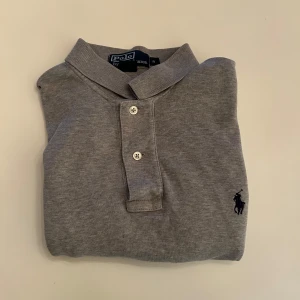 Grå pikétröja från Polo Ralph Lauren - Snygg grå pikétröja från Polo Ralph Lauren i 100% bomull. Klassisk design med korta ärmar, krage och två knappar framtill. Diskret broderad logga på bröstet. Perfekt för en clean och stilren look.