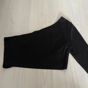Svart one shoulder topp från SHEIN - Säljer en svart, tight one shoulder topp från SHEIN. Toppen har lång ärm på ena sidan och är axelbandslös på andra, vilket ger en snygg asymmetrisk look. Gjord i ett tunt stretchigt material som sitter skönt mot kroppen.