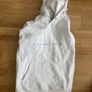 Vit hoodie från Jack & Jones - Snygg vit hoodie från Jack & Jones med minimalistisk svart logga på bröstet. Klassisk känguruficka framtill och huva med snörning. Perfekt för en chill och stilren look.