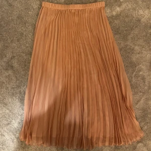 Plisserad beige långkjol - Snygg plisserad långkjol i en mjuk beige ton. Kjolen har ett flowigt fall och resår i midjan för extra komfort. Perfekt för dig som gillar en stilren och trendig look med mycket rörelse i tyget.