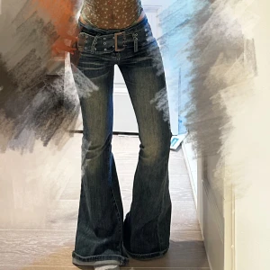 Blå los Waist jeans med bredt bälte - Säljer ett par blå bootcut jeans med slitningar och coolt tvättat mönster. Byxorna har låg midja och ett brett bälte med stora nitar och metallspänne. Jeansen är i klassisk denim och har en riktigt snygg 00-talsvibe.