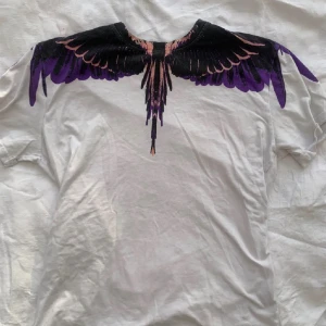 Vit Marcelo burlon t-shirt med lila och svart vingmönster - Säljer en vit Marcelo burlon t-shirt med ett unikt vingmönster i svart och lila över axlar och hals. T-shirten har rund halsringning och korta ärmar och så är inte priset fast och kan diskuteras vid snabb affär. Perfekt för dig som vill sticka ut med en cool design!