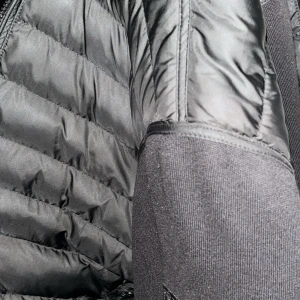 Svart pufferjacka med stickade partier - Säljer en svart pufferjacka med snygga stickade partier på sidorna och ärmarna. Jackan har quiltade paneler och en modern look som passar perfekt till kalla dagar. Dragkedja framtill och stilren design.