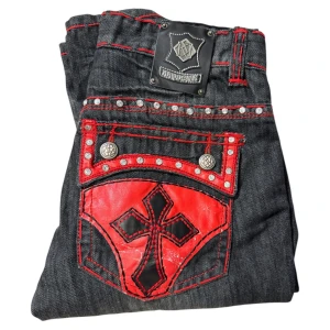 Svarta jeansbyxor med röda detaljer från Kayden.k - Unika svarta jeans med röda kontrastsömmar, stora bakfickor med röda och svarta korsapplikationer samt nitar. Märkespatch i läder baktill och dekorativa knappar. Snyggt slitna detaljer framtill och bak. Lite nötta på bakfickorna o coin pocket men inget man tänker på.
