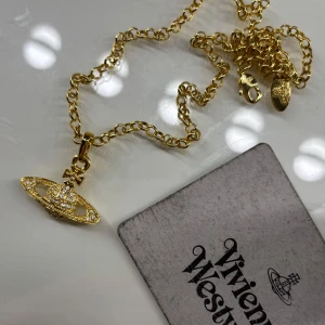 Guldigt halsband med orb-hänge från Vivienne Westwood - Säljer ett ikoniskt halsband från Vivienne Westwood i guldton med det klassiska orb-hänget dekorerat med gnistrande stenar. Kedjan är grov och har märkets logga ingraverad på brickan vid låset. Perfekt statement-piece!