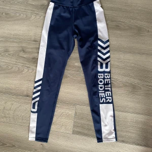Mörkblå leggings från Better Bodies - Snygga mörkblå leggings från Better Bodies med vita sidopaneler och logga längs benet. Tight passform och hög midja, perfekta för gymmet eller annan träning. Materialet är stretchigt och bekvämt.
