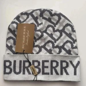 Gråvit mössa från Burberry med hjärtmönster - Snygg gråvit mössa från Burberry med stort logotyptryck framtill och stickat hjärtmönster i mörkgrått. Mössan har uppvikt kant och är tillverkad i mjukt stickat material. Perfekt för att ge din outfit en exklusiv touch.