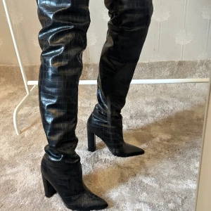 Svarta overknee stövlar från PLT - Säljer ett par svarta overknee stövlar från Prettylittlething. Har höga klackar och spetsig tå,  krokodilpräglat mönster. Klacken är ca. 11 cm och skorna kommer lite över knäna (se bild). Använda några gånger och har lite skråmor framtill på skon (syns på bild). 