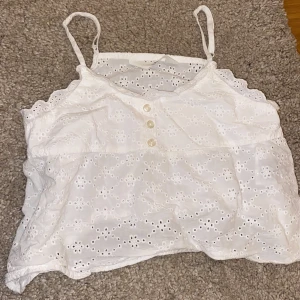 Vit broderad linnetopp från H&M - Säljer en söt vit linnetopp från H&M med broderade blommor och tre dekorativa knappar framtill. Toppen har smala axelband och är croppad med fina spetsdetaljer längs kanterna. Perfekt för varma dagar!