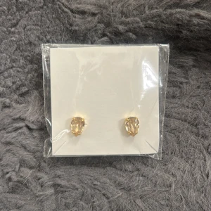Guldiga örhängen med stenar från Caroline Svedbom - Säljer ett par eleganta örhängen från Caroline Svedbom. Örhängena är guldiga med droppformade, ljusa stenar som ger en lyxig känsla. Perfekta för att lyfta vilken outfit som helst. Endast öppnade en gång aldrig använda. I färgen light silk. 