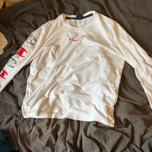 Vit långärmad tröja från Champion - Säljer en vit långärmad tröja från Champion med tryckta loggor i svart och rött längs ena ärmen och liten röd Champion-text på bröstet. Perfekt för en avslappnad och sportig stil.