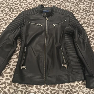 Svart skinnjacka från Cordina - Säljer en svart skinnjacka från Cordina i storlek M. Jackan har snygga quiltade detaljer på axlar och ärmar, dragkedjor framtill och flera fickor med dragkedja. Perfekt för dig som gillar en stilren och edgy look.