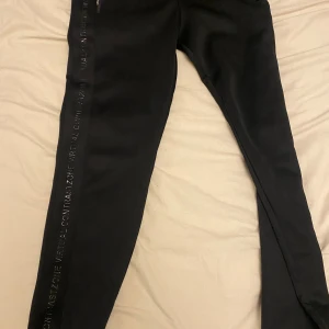 Svarta tighta träningsbyxor med textdetalj - Svarta träningsbyxor med tight passform och diskret text längs ena benet. Byxorna har dragkedja upptill och är perfekta för gymmet eller löpning. Snygg och sportig look som passar till många olika träningspass.