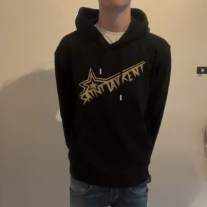 Saint Laurent hoodie - Säljer en svart hoodie från Saint Laurent med ett tryck i guld på framsidan. Strl S. Den är inte så använd, så skulle kunna säga att skicket är 9/10