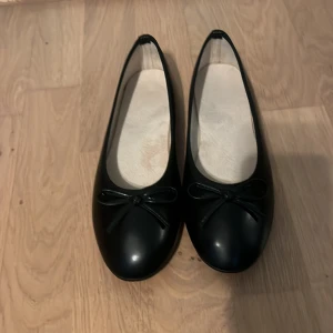 Svarta ballerinaskor med rosett - Säljer ett par klassiska svarta ballerinaskor med rund tå och en liten rosett framtill. Skorna har en enkel och stilren design som passar till många olika outfits. Perfekta för dig som gillar en tidlös look. Dem är helt nya också!
