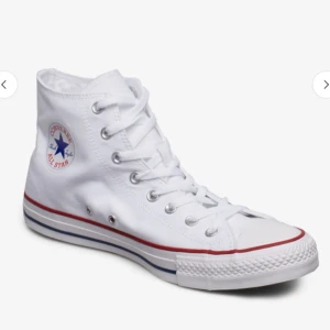 Converse - Klassiska vita höga Converse All Star sneakers med snörning och rund tå. Skon har en vit canvasöverdel, röd och blå rand längs sulan samt Converse-logga på sidan. Perfekt för en avslappnad och tidlös stil. 