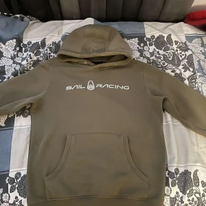 Olivgrön hoodie från Sail Racing - Säljer en olivgrön hoodie från Sail Racing med vit logga på bröstet. Tröjan har huva, magficka och ribbade muddar. Perfekt för en avslappnad stil.