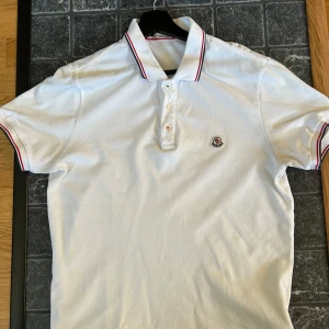 Moncler pike vit  - Vit piké t-shirt från Moncler med klassisk krage och knäppning framtill. Kragen har röda och blå ränder och Moncler-logga är broderad på bröstet. Piken är i storlek medium, piken är i bra skick förutom tre små hål man kan se på sista bilden inget man märker när man har på sig piken. Om ni har några frågor är det bara att skriva 