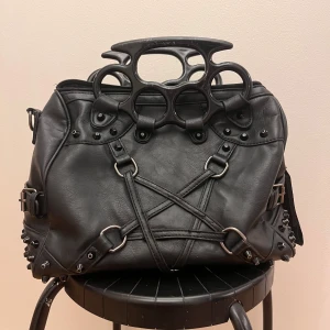 Svart axelväska med unikt handtag - Cool svart väska i skinnimitation med ett unikt handtag som ser ut som knogjärn. Väskan har flera metalldetaljer, dragkedjor och man kan även sätta på ett längre band på väskan. Den har både fack i väskan och utanpå på baksidan. Dock en defekt där sömmen har spruckit upp vid dragkedjan upptill. Mått.. Bredd 30cm, Höjd 24cm, Djup 16cm😸💞 