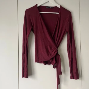 Vinröd omlottopp från 157 - Säljer en vinröd omlottopp från 157 i mjukt material. Toppen har långa ärmar och knytdetalj i midjan som ger en snygg passform. Perfekt att styla med jeans eller kjol för en trendig look!