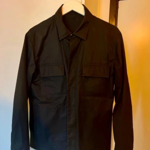 Tiger Of Sweden - Svart overshirt med fickor - Stilren svart overshirt med klassisk krage och två bröstfickor med lock. Jackan har knäppning framtill och raka ärmar. Perfekt för lager-på-lager och enkel att matcha med olika outfits. Jackan är använd under en sommar, så i väldigt bra skick.  Köptes i Göteborg för 1999kr.  Overshirt från Tiger of Sweden Jeans. CHILLZ SHIRT. Tygnummer: W69911 Färgkod: 096 - Normal passform - Klassisk krage - Fyrkantig manschett - Dold knappknäppning - Öppna sidofickor - Två bröstfickor med lock och knapp
