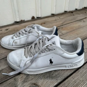 Vita sneakers från Polo Ralph Lauren - Säljer ett par klassiska vita sneakers från Polo Ralph Lauren med mörkblå detaljer och logga på sidan. Skorna har snörning och rund tå, perfekt för en stilren vardagslook. Tvättas innan den skickas inga hål i 