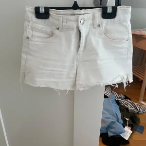 Vita jeansshorts med fransar - Säljer ett par lowwaist vita jeansshorts med råa franskantsdetaljer och klassisk femficksdesign. Shortsen har knapp- och dragkedjestängning samt bälteshällor. Perfekta för varma dagar och enkel att matcha med olika toppar.